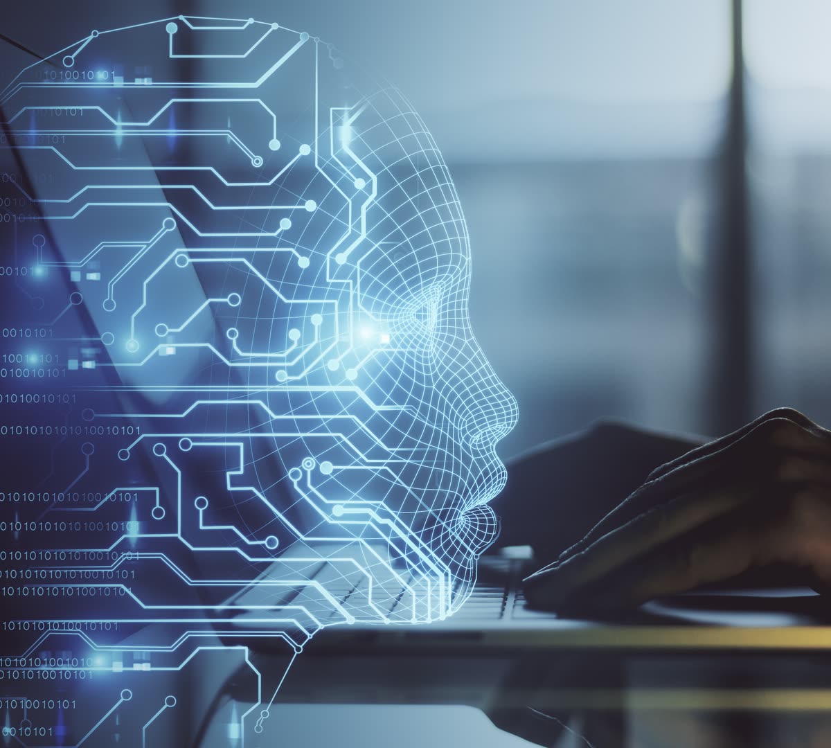 A glowing wireframe human head in profile, drawn from circuit traces and streams of binary digits, over a photograph of hands typing at a laptop keyboard.
